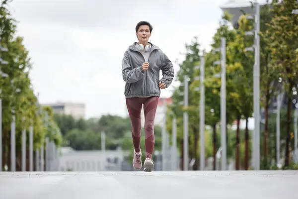 stock image Full shot of fit elderly woman in sportswear jogging along wide walkway in morning city park working out on regular basis, copy space