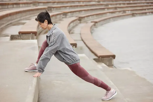 stock image Side view of fit senior woman in pink sportswear stretching leg muscles on huge concrete steps of amphitheater getting ready to run jogging outside in city park, copy space