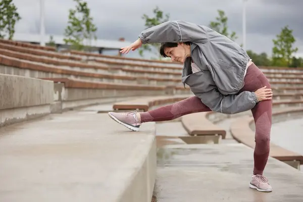 stock image Side view of elderly woman in pink leggings stretching back muscles side bending over leg on huge concrete steps of amphitheater working out regularly in city park, copy space