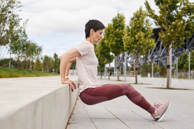 Pembe taytlı yaşlı bir kadının kenar manzarası. Şehir parkında günlük antrenman için triseptik bank dalışı yapıyor.