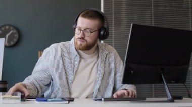 Ofiste bilgisayar başında çalışırken kulaklık takan genç beyaz programcı akıllı telefondan bildirimi kontrol ediyor.