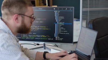 Gözlük takan genç sakallı programcı IT şirketinin ofisinde dizüstü bilgisayarda daktilo kodunu kullanıyor.
