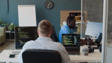 Modern ofiste siber güvenlik kodları üzerinde çalışan tanınmayan beyaz bir adamın arka görüşü, siyah meslektaşı ona dijital tablet veriyor.
