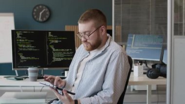 Genç beyaz programcının orta boy portresi modern ofiste iş yerinde, dijital tablet kullanarak kameraya bakıyor.