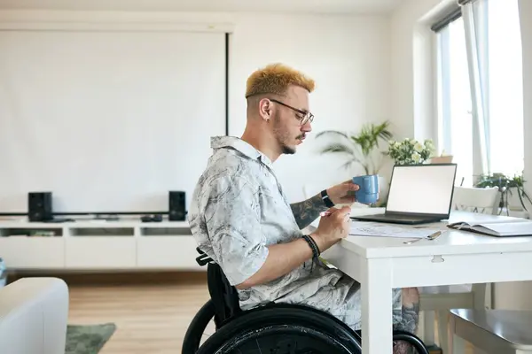 stock image Man in wheelchair working from home, sitting at desk with laptop and holding blue coffee cup, facing left side, next to large window and white bookshelf, wearing casual clothes