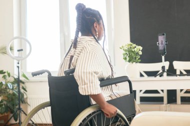 Tekerlekli sandalyede oturan kadın bilgisayar ekranına bakıyor. Ofiste bitki ve halka ışığıyla sanal bir toplantıya katılıyor.