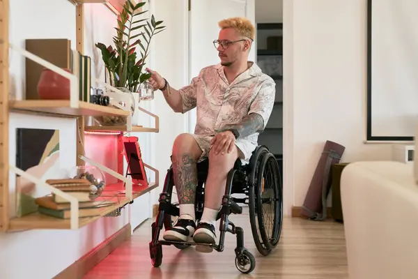 stock image Caucasian man in wheelchair moving through modern apartment reaching for items on shelf. Living room with wooden floor and stylish decor creating a welcoming environment