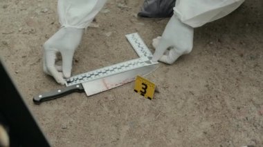 Koruyucu elbise giyen, yerde kanlı bıçağı inceleyen ve dışarıdaki suç mahallini araştırırken cinayet aleti olarak kanıt torbasına koyan tanınmamış adli tıp uzmanının yakın çekimi.