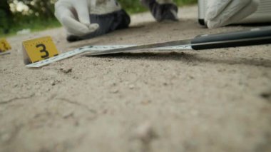 Koruyucu giysi üzerine adli tıp uzmanının cinayet aletini bulup eldivenli ellerle plastik kanıt torbasına koyduğu görüntüler.