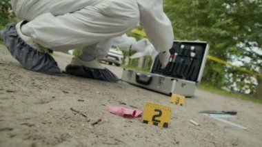 Koruyucu giysi konusunda tanınmayan adli tıp uzmanının plastik poşette kanıt toplayıp cinayet mahalline koyduğu düşük açılı bir çekim.