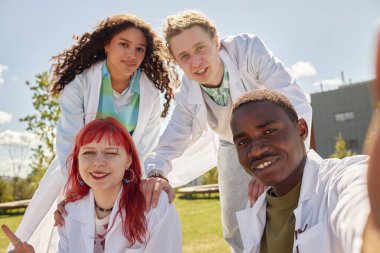 Farklı etnik kökenlere sahip dört gülümseyen tıp öğrencisinin portresi kampüste birlikte takılırken selfie çekiyor.