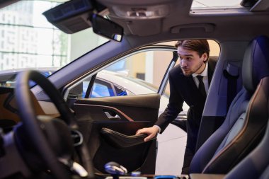 Asyalı erkek otomobil satıcısı yeni arabanın kapısını açıyor. Aracın içini inceliyor. Araba galerisinde deri döşeme var.