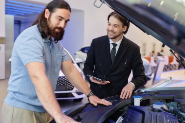 Orta boy gülümseyen siyah takım elbiseli Orta Asyalı otomobil satıcısı araba alıcısına araba teknik özellikleri konusunda tavsiye veriyor, galeri merkezindeki yeni aracın kaputunun altına bakıyor.