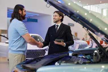 Orta boy gülümseyen Orta Asyalı otomobil satıcısı yetişkin bir adamla el sıkışıyor ve bayilik merkezinde yeni bir araba alıyor.