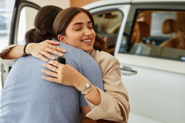 Galeri merkezinden başarılı araba alımlarından sonra erkek arkadaşını kucaklayan mutlu genç bir kadının orta boy fotoğrafı.