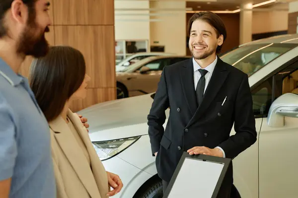 stock image Medium shot of Central Asian male auto dealer talking to couple planning to buy new car. Smiling motor trader assisting clients at dealership center