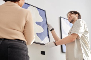 İki gülümseyen sanat galerisi işçisinin düşük açılı görüntüsü müzedeki duvara dikkatlice soyut sanat tablosu asıyor.