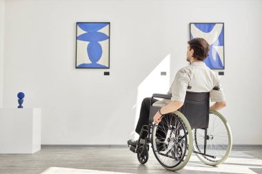 Tekerlekli sandalye kullanan bir adamın erişilebilir sanat galerilerinde ya da müzede güneş ışığı fotokopi alanı ile aydınlatılmış resimlere bakışıyla tam boy görünümü