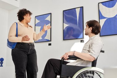 Yandan bakınca gülümseyen siyahi kadın, tekerlekli sandalye kullanan adamla konuşuyor modern sanat galerisinde soyut tabloları tartışıyor.