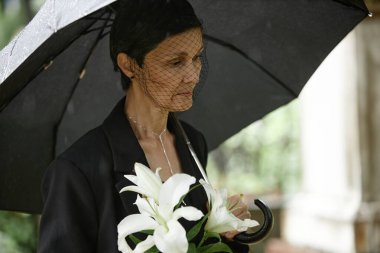 Side view of senior woman under black umbrella wearing mourning veil and grieving loss of departed loved one at grave while holding white lilies at cemetery, copy space clipart