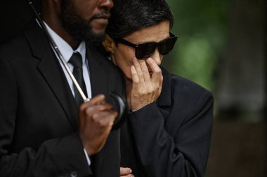 Ağlayan, siyah güneş gözlüklü yaşlı bir kadının, kocasının omzuna sarılıp gözyaşlarını silerken, mezarlıktaki anma töreninde kaybettiği yas sonrası teselli ararken orta boy fotoğrafı.