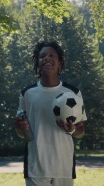 Orta boy, neşeli Afrikalı Amerikalı genç futbolcunun takım arkadaşıyla sohbet ederken güneşli yeşil park boyunca birlikte futbol antrenmanına giderken çekilmiş fotoğrafı.