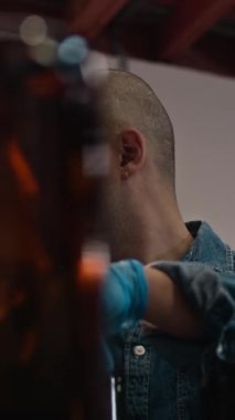 Kafası tıraşlı, kot gömlek giyen ve bira fabrikasında çalışırken şişeleme işlemine katılan, endüstriyel nitril eldivenli beyaz bir adam.