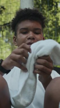 Afrika kökenli Amerikalı genç futbolcunun açık hava eğitiminden sonra yeşil çimlerde dinlenirken beyaz havluyu omuzlarına giydiği orta boy bir fotoğraf.