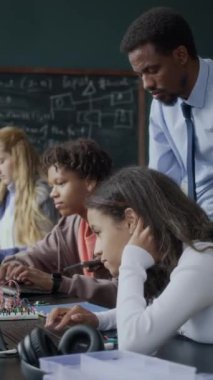 Genç Afrikalı Amerikalı erkek öğretmenin kodlama ve elektronik dersinde çoklu etnik kökenli genç öğrencilere yardım ettiği dikey çekim.