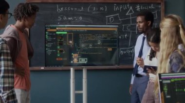 Genç Afro-Amerikan erkek programlama öğretmeni ve çok ırklı genç kız öğrencilerin ekrandaki yazılım arayüzüne bakarken görüntüsü.