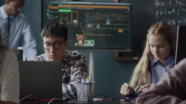 Modern sınıftaki yerlerinde oturan çok ırklı genç öğrenciler programlama ve mühendislik dersleri sırasında dizüstü bilgisayar ve devre kartı montajı yapıyorlar.