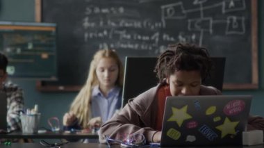 Orta boy Afrika kökenli Amerikalı erkek programlama öğretmeni grup dersi sırasında dizüstü bilgisayarın önünde oturan genç çocuğa yardım ederken.