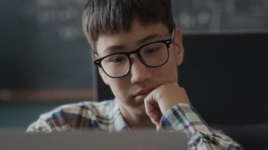 Gözlüklü Asyalı gencin, sınıfta programlama yaparken dizüstü bilgisayara ilgiyle bakarken orta ölçekli yakın çekimi.