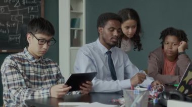 Afrika kökenli Amerikalı erkek öğretmenin programlama ve mühendislik dersi sırasında çok ırklı z öğrencilerine kablolarla devre kartı gösterirken orta boy fotoğrafı.