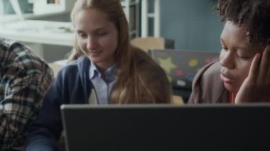 Dizüstü bilgisayarın önünde oturan ve modern sınıftaki programlama dersleri sırasında sohbet eden etnik çeşitlilikte genç öğrencilerin belini kaldırın.