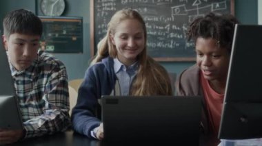 Modern programlama sınıfında, bilgisayarlarının başında oturmuş sohbet eden üç farklı etnik gruptan öğrencinin orta boy fotoğrafı.