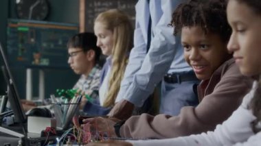 Modern mühendislik dersinde siyahi erkek öğretmen onlara yardım ederken elektronik projesinde çalışan etnik çeşitlilikte genç erkek ve kız öğrenciler sohbet ediyorlar ve daktilo yazıyorlar.