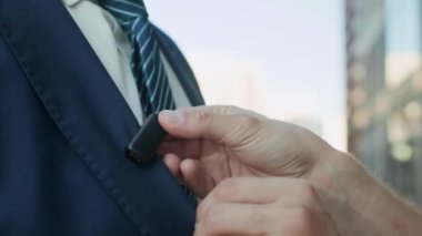 Resmi mavi takım elbiseli, beyaz gömlekli ve mavi çizgili kravatlı, şehir ortamında kayıt yapmadan önce iki kablosuz mikrofona sahip tanımlanamayan erkek haber sunucusunun yakın çekimi.