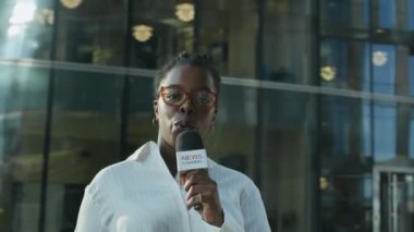 Gözlüklü ve beyaz çizgili tişörtlü Afro-Amerikan kadın gazetecinin şehrin göbeğinde güneşli bir sokakta dikilirken mikrofonla haber yaptığı orta ölçekli bir fotoğraf.