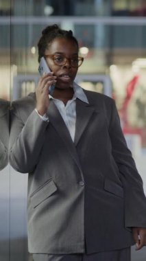 Gri takım elbiseli başarılı Afro-Amerikan iş kadınının dikey görüntüsü. Açık havada akıllı telefondan arama yapıyor. Bulanık şehir manzarası arka planında duruyor.