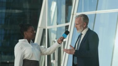 Orta boy, beyaz takım elbiseli, beyaz gömlekli ve kravatlı bir iş adamı Afro-Amerikan kadın TV gazetecisine röportaj yaparken, güneşli caddede cam bina karşısında dururken.