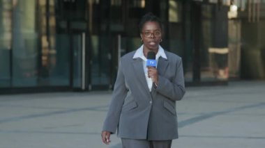 Gri takım elbiseli, kendine güvenen genç siyahi kadın muhabirin şehir merkezinde haberleri izlerken mikrofonla kameraya doğru yürürken, arka planda bulanık cam binayla görüntüsü.