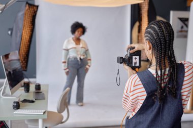 Afrika kökenli Amerikalı genç bir kadının profesyonel fotoğraf stüdyosundaki beyaz arka planda kadın modelin fotoğraflarını çekmesi.