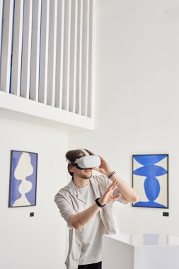 Müzede VR kulaklık takan ve podyumdaki görünmez dijital sanat eserleriyle etkileşime giren bir adamın minimum dikey portresi.