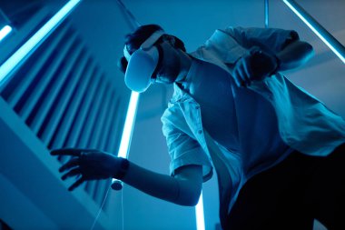 Modern sanat fuarında VR kulaklık takan bir adamın düşük açılı portresi. Mavi ışıklar fütüristik dijital siber uzayın keyfini çıkarıyor.