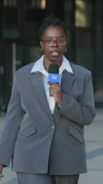 Gri takım elbiseli, kendine güvenen genç siyahi kadın muhabirin şehir merkezindeki haberleri izlerken mikrofonla kameraya doğru yürüdüğü ve arka planda bulanık cam binalar oluşturduğu dikey bir görüntü.