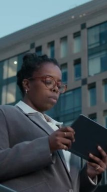 Akşamları modern cam ofis binalarının önünde dijital tablet kullanarak merdivenlerden inerken zarif gri takım elbiseli ve gözlüklü başarılı Afrikalı iş kadınının dikey orta pozu.