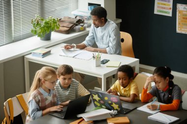 Modern sınıftaki büyük bir masada ders çalışırken çift olarak çalışan ilkokul öğrencilerinin yüksek açılı görüntüleri.