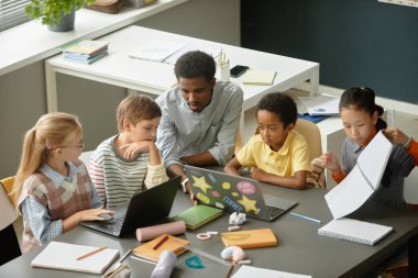 Afrika kökenli Amerikalı erkek öğretmenin çocuklara laptopları kullanmada yardım eden ve modern sınıflarda ödevlerini açıklayan bir grup çoklu etnik kökenli genç öğrenciyle aynı masada yüksek açılı fotoğrafı.