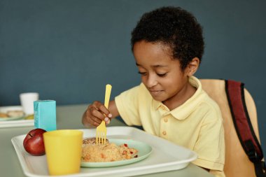Orta boy Afro-Amerikan çocuk masada oturmuş okul kafeteryasında lezzetli yemekler yerken, fotokopi çekerken.